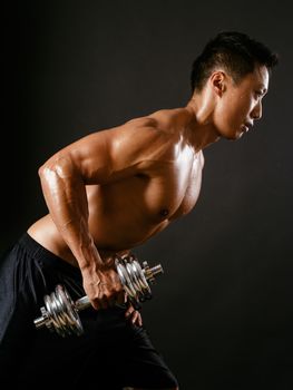 Photo of an Asian male exercising with dumbbells and working his triceps over dark background. Focus is on the arm.