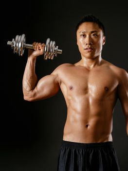 Photo of an Asian male exercising with dumbbells and doing a single shoulder press over dark background.