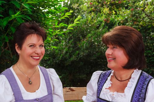 Portrait of two senior women from Bavaria, who sit together in a dirndl on a bench in the garden and enjoy your free time