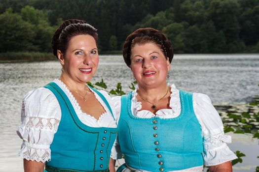 Two happy Bavarian ladies in dirndls standing side by side in front of a lake outdoors, upper body portrait