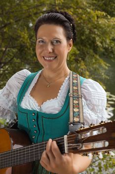 Smiling middle-aged woman in Bavarian Dirndl and braided her hair stands on the lake and playing guitar