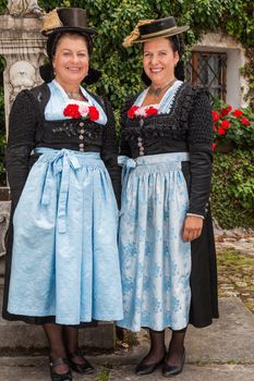 Two charming ladies in traditional dirndls with festive red flowers tucked into the bodices of the dresses standing together outdoors