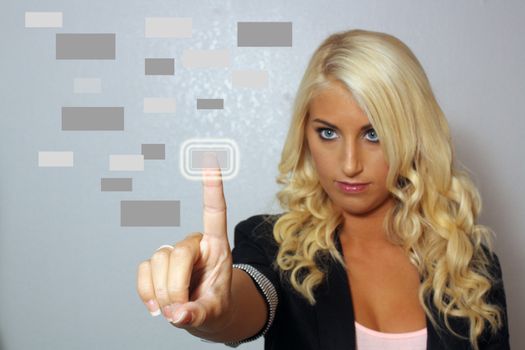 An extraordinarily beautiful young blonde isolated on a textured grey background pushes a futuristic virtual button.  Selective focus on her finger.