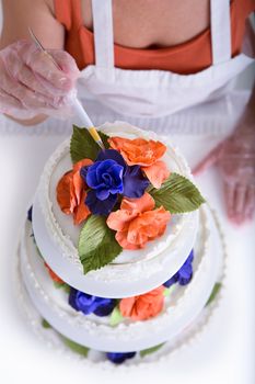 Woman doing latest touches to a cake with her gloves on, shot from above, cake have orange and purple flowers with green leaves