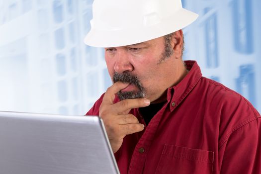 Hardhat worker perplexed, working on the tough issues with his laptop, he has a serious thoughtful look that he values his job, he is wearing red shirt and isolated on bluish background, copy space on hardhat and laptop