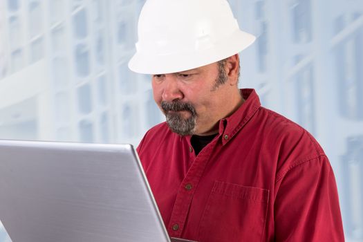 Hardhat worker working on the tough issues with his laptop, he has a serious thoughtful look that he values his job, he is wearing red shirt and isolated on bluish background, copy space on hardhat and laptop