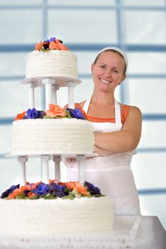 Happy baker lady happily smiling large infront of her cake with her apron and white bandanna, cake has fondant ruffles on the side and decorated with orange and purple gum paste roses