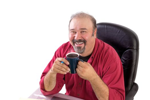 Middle age man smiling happily to camera with his morning coffee at his desk, wearing red shirt and sitting on his black leather chair