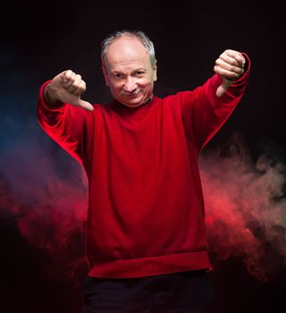 An elderly man in a red sweater shows thumbs down on a dark background