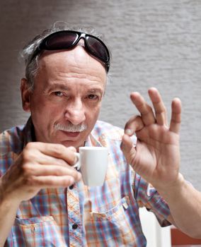 Portrait of smart old man drinking cup of coffee and showing sign OK with fingers