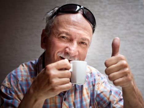 Portrait of smart old man drinking cup of coffee and showing sign OK with fingers