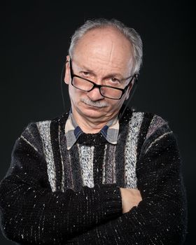 An elderly man with glasses and crossed arms looks skeptically on a dark background