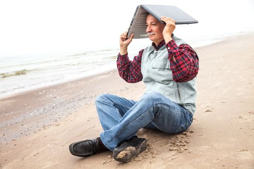 Old man sitting on a sea beach and holding a laptop on his head on a foggy day