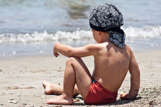 Little boy sitting on a beach looking on the waves