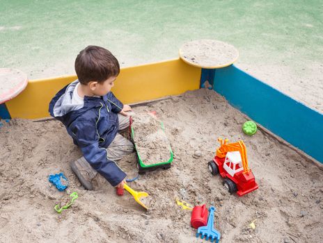 Cute boy in jacket playing in sandbox with a lot of sand toys