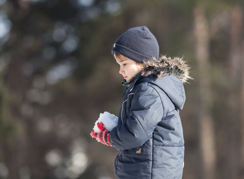 Cute boy in a winter jacket, playing with snow on blurred nature background