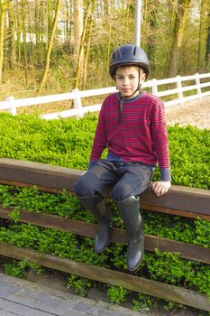 cute teenage  boy in helmet and boots for horse riding sitting next to manege