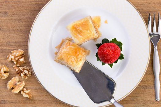 Couple pieces of home made baklava getting ready to be served with stainless-steel spatula on old bamboo cutting board along with walnuts and strawberry