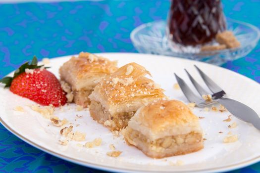 Three pieces of golden color baklava garnished with walnuts and strawberry and served on oval shape plate on green and blue color table cloth