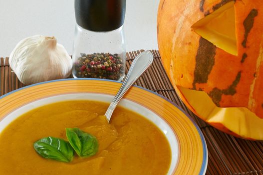 A bowl of pumpkin soup with a couple of basil leaves, pepper, garlic and a Halloween pumpkin