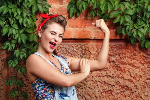 Beautiful pin-up girl in denim overalls and a red bandana  winks and demanstriruet bicep