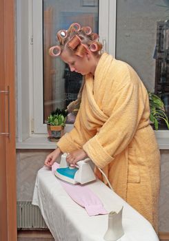 Girl with hair curlers in a dressing gown irons clothes.