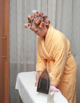 Girl with hair curlers in a dressing gown irons clothes.