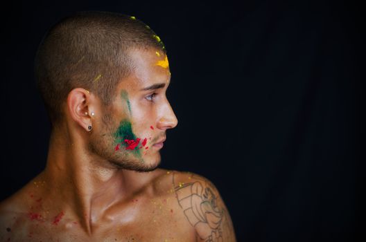 Attractive young man shirtless, skin painted all over with bright colors looking to a side, profile view