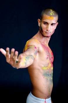 Handsome young man with skin all painted with colors, arm stretched towards camera on dark background