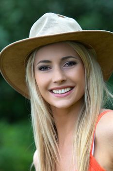 Beautiful smiling woman with long straight blond hat wearing a trendy wide brimmed hat outdoors in the park and giving the camera a toothy smile