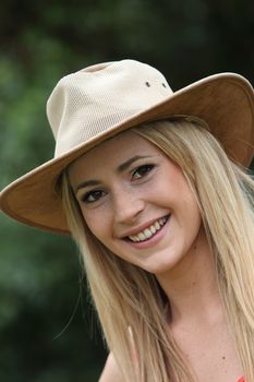Happy young blond woman with a beaming smile standing in a lush garden in a trendy hat