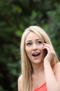 Beautiful young blond woman reacting in awe to a call on her mobile phone standing with her mouth open in astonishment