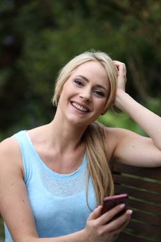 Laughing beautiful woman with a blond ponytail sitting on a wooden garden bench with her mobile phone in her hand