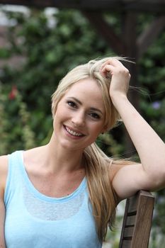 Pretty blond woman with a beautiful smile and her hair in a casual ponytail leaning on a wooden garden chair looking at the camera