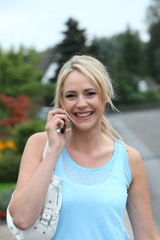 Happy woman with long blond hair tied back in a ponytail chatting on her mobile while standing in a road with her handbag over her shoulder