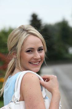 Smiling casual young blond woman with her hair in a ponytail and a handbag over her shoulder turning to smile at the camera