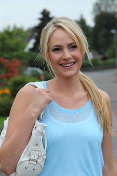 Happy young woman in casual summer clothes standing outdoors in the garden with a handbag over her shoulder smiling at the camera