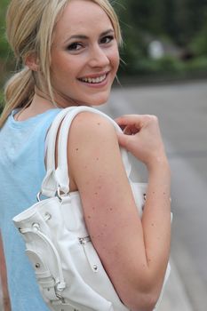 Pretty young blond woman with a handbag over her shoulder turning to look back at the camera with a delightful smile