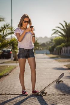 Beautiful young girl with a skateboard using her smartphone outdoor on summer