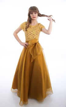 Teen girl stands against a white, isolated background while wearing a beautiful gold colored formal prom gown twirling her hair