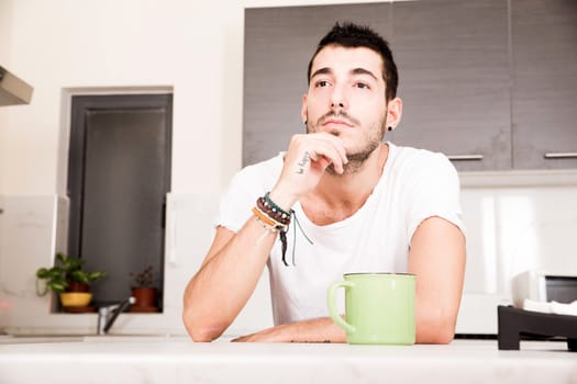 A young man sitting in the kitchen and drinking coffee.