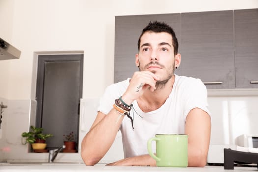 A young man sitting in the kitchen and drinking coffee.