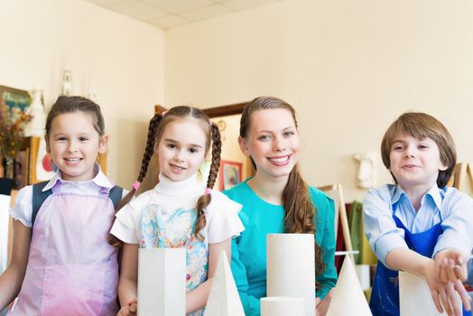 children with the teacher standing at the table, smiling and looking into the cam