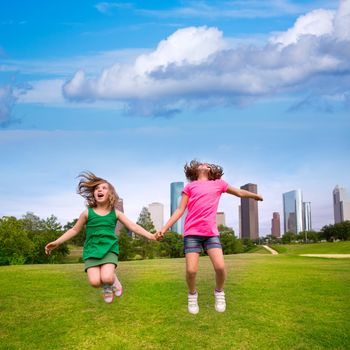 Two sister girls friends jumping happy holding hand in urban modern skyline on park lawn