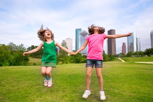 Two sister girls friends jumping happy holding hand in urban modern skyline on park lawn