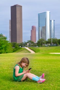 Blond kid girl playing with smartphone sitting on park lawn in city skyline background