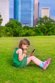 Blond kid girl playing with smartphone sitting on park lawn in city skyline background