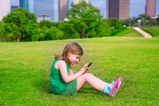 Blond kid girl playing with smartphone sitting on park lawn in city skyline background