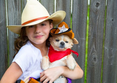 Beautiful cowboy kid girl holding chihuahua dog with sheriff hat in backyard wooden fence