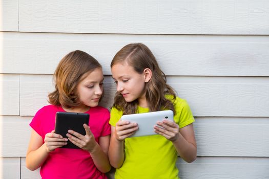 Twin sister girls playing with tablet pc happy on white wall looking each other